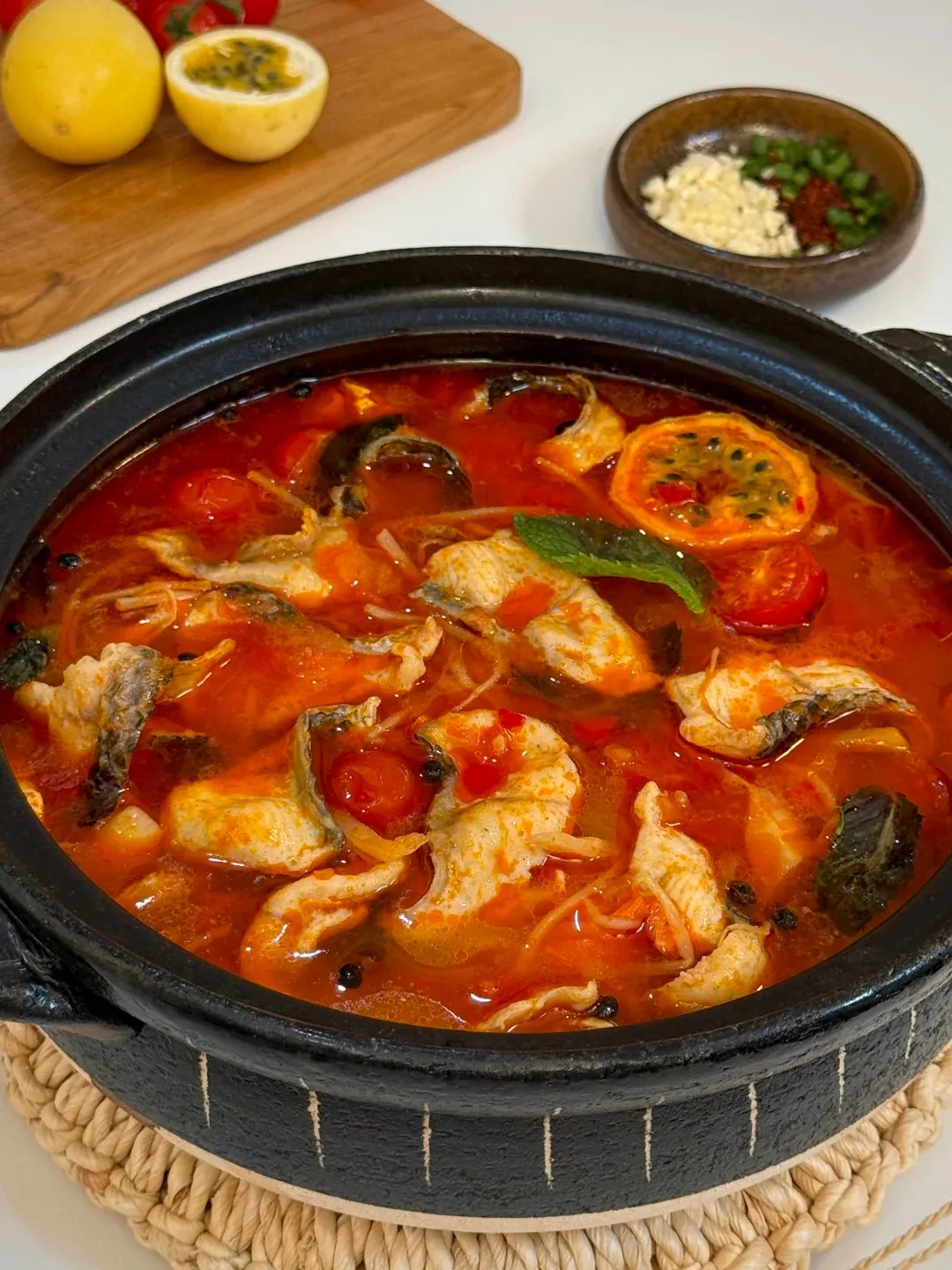 A close-up of a black, rustic hot pot filled with a vibrant red, spicy broth containing pieces of white fish, cherry tomatoes, black peppercorns, and a slice of passion fruit. The pot sits on a woven placemat, with a wooden cutting board holding two lemons and a halved passion fruit visible in the upper left corner, and a small bowl of chopped garlic and chili flakes in the upper right.