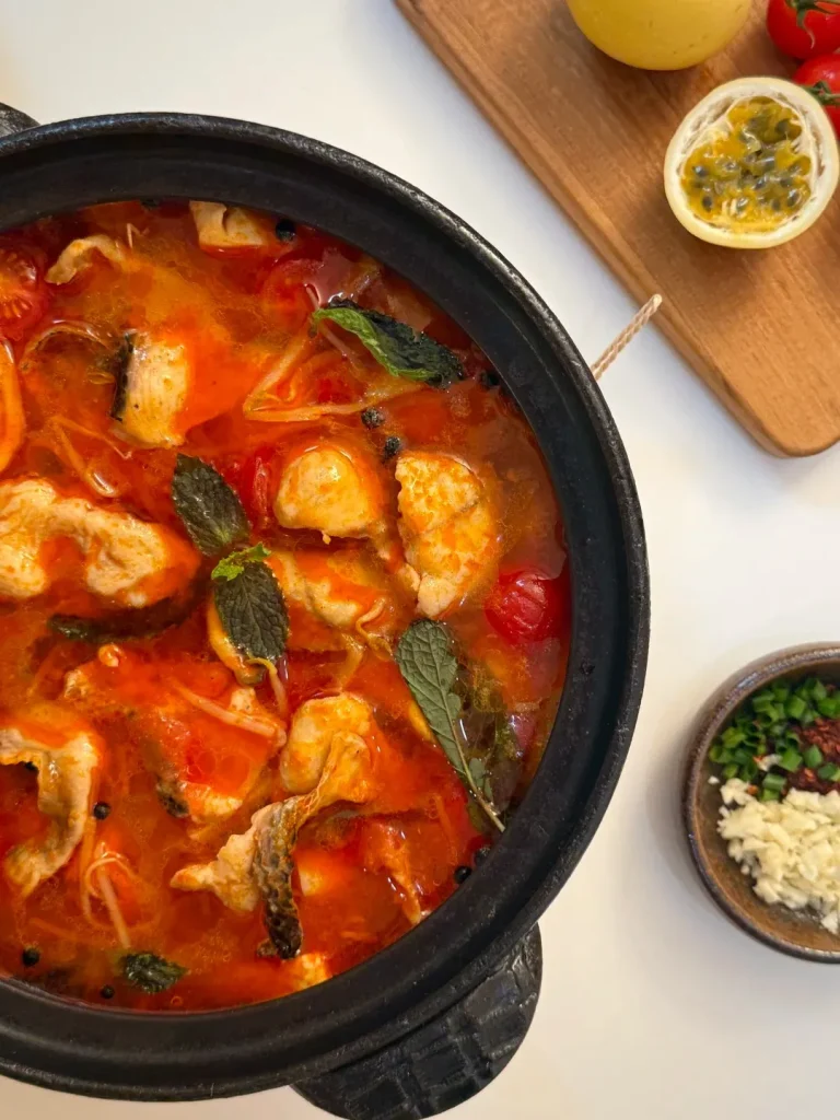 A close-up, overhead view of a black pot filled with a vibrant red and orange sour fish soup, featuring chunks of white fish, cherry tomatoes, and fresh mint leaves. To the right, a small ceramic bowl contains minced garlic, chopped green onions, and chili flakes. In the background, a wooden cutting board holds a halved passion fruit and two cherry tomatoes.
