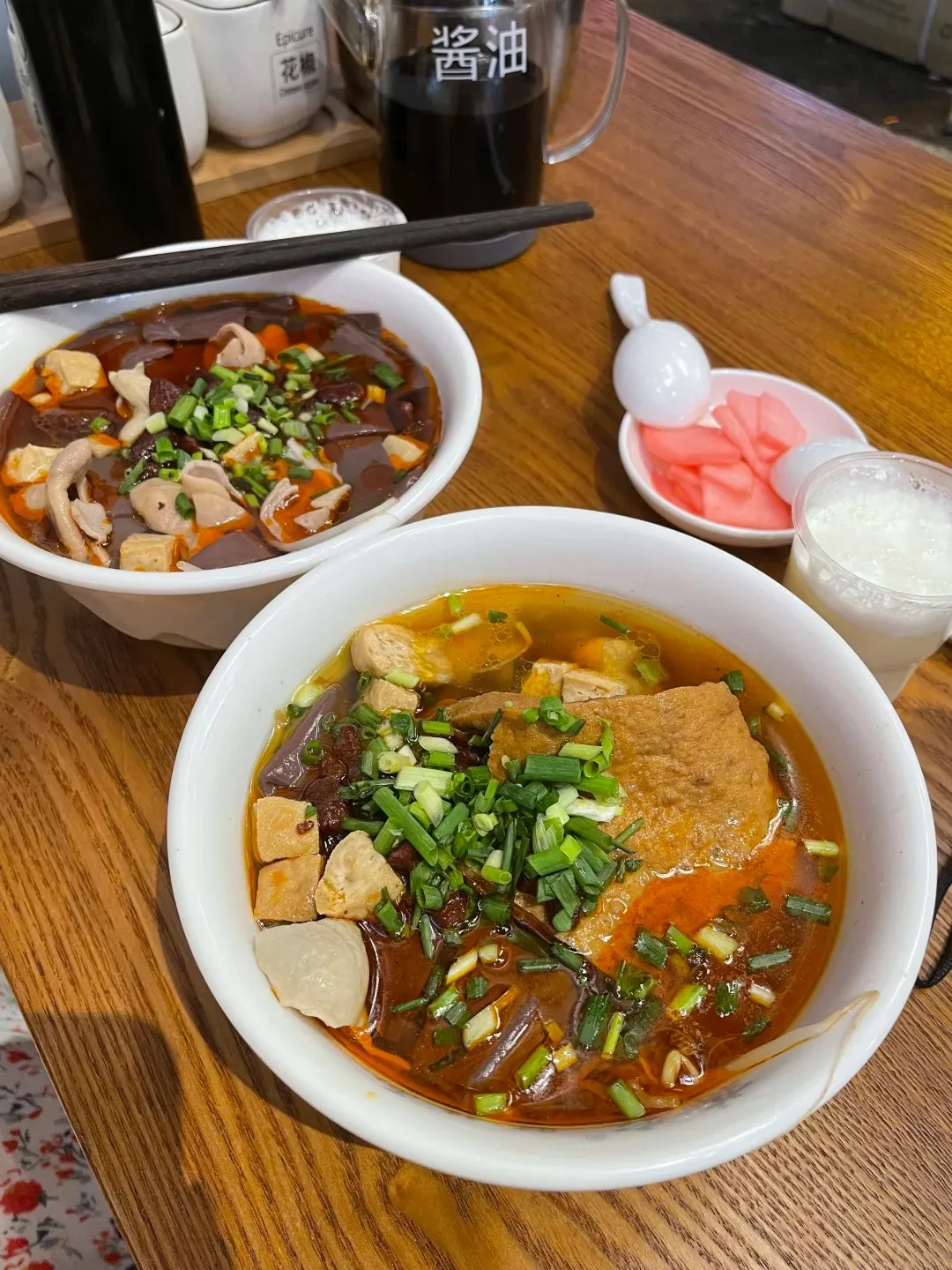 Two white bowls of steaming spicy and sour noodle soup filled with dark broth, tripe, blood, tofu, meat, and chopped green onions, with chopsticks resting on the edge of the bowl in the background. A small white dish of pickled pink ginger and a clear cup of white drink are to the right.