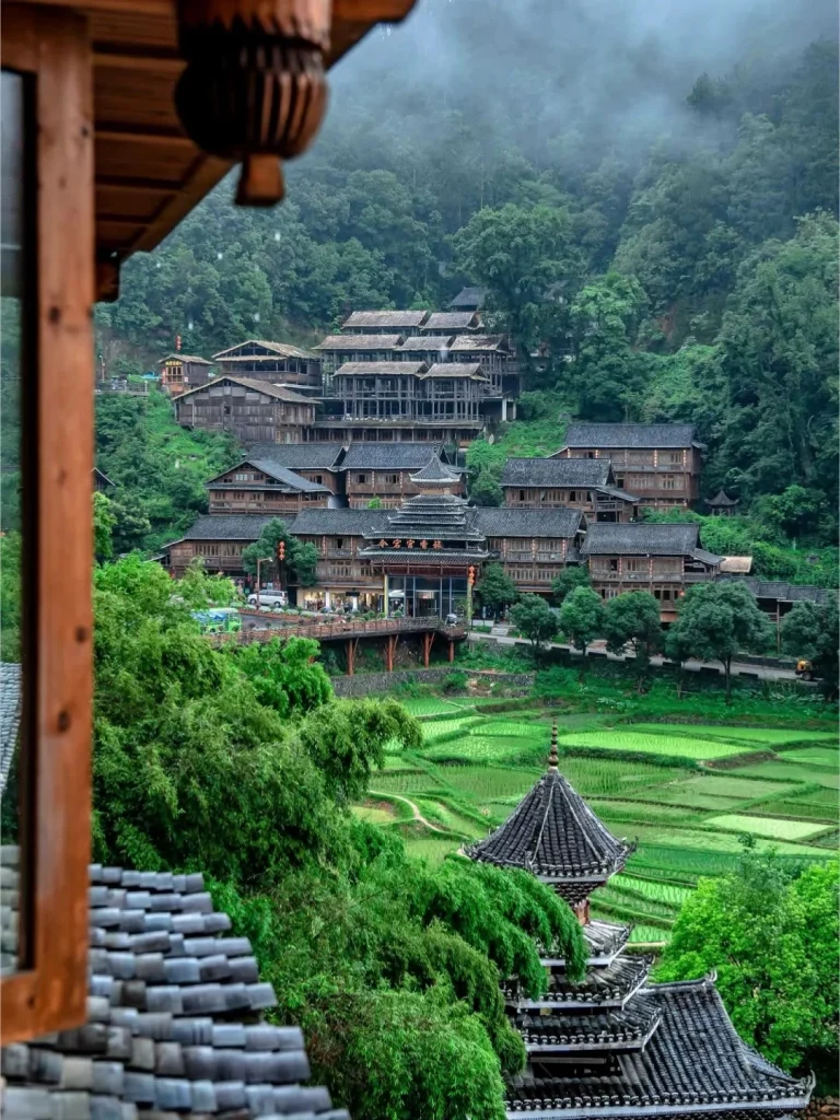View from a wooden balcony overlooking a misty, mountainous landscape. In the mid-ground, traditional wooden buildings with tiled roofs are built on a hillside. In the foreground, lush green trees frame tiered, vibrant green rice terraces. A multi-tiered pagoda with a dark, curved roof sits in the mid-right foreground.
