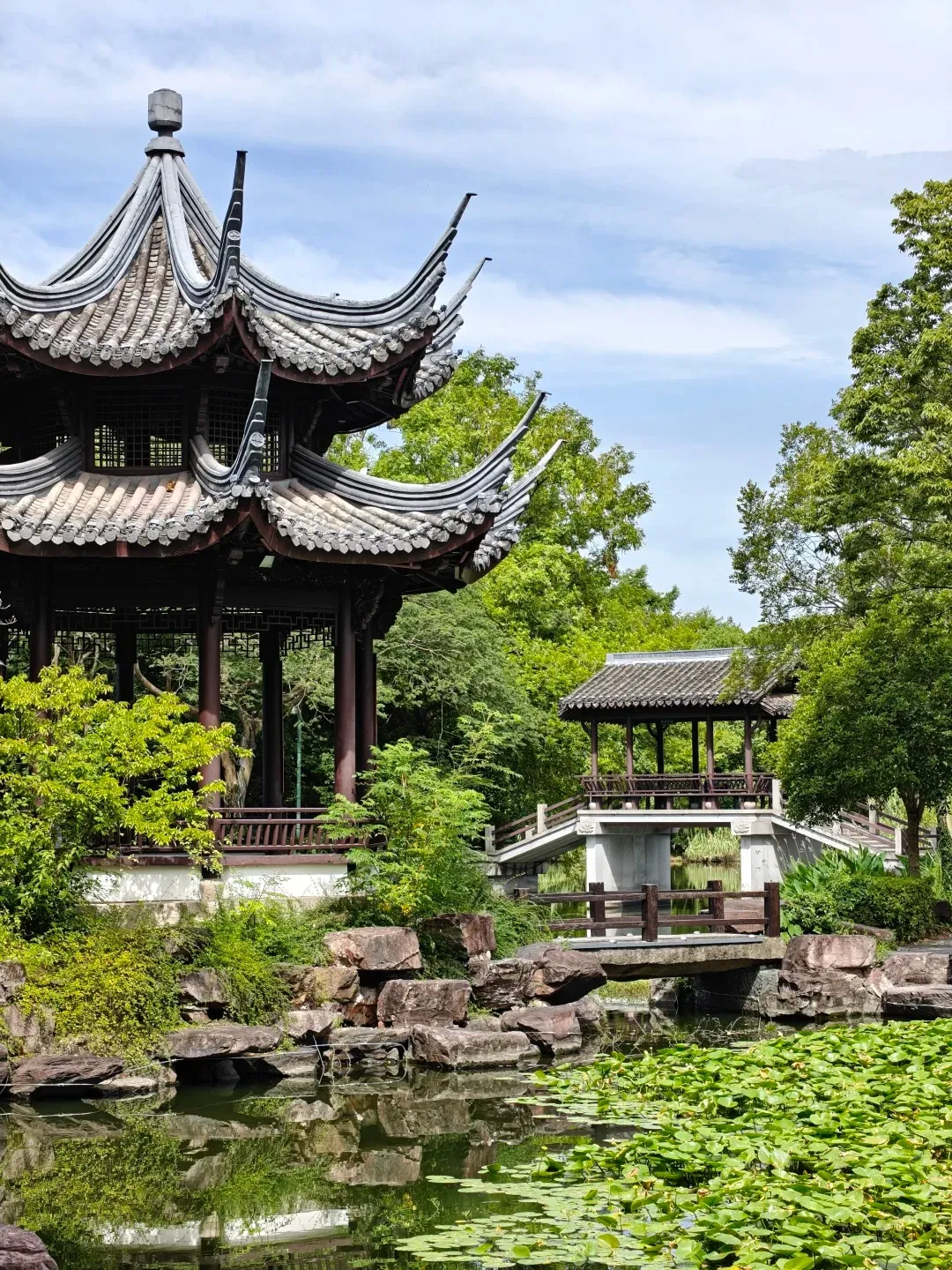 A traditional Chinese pagoda with multiple tiered, curved roofs stands to the left, partially obscured by lush green trees. In the center and right, a stone bridge crosses a tranquil pond filled with lily pads, leading to a smaller, covered pavilion on a raised platform. Rocks and dense greenery surround the water, and the sky above is partly cloudy.