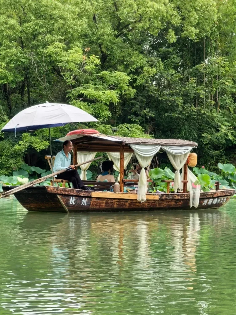 A man with short dark hair wearing a light blue shirt and black pants rows a wooden boat with a canopy and white curtains. There are two passengers seated in the boat. The boat is on a body of green water, with tall green trees and foliage in the background. Lotus pads are visible to the left of the boat.
