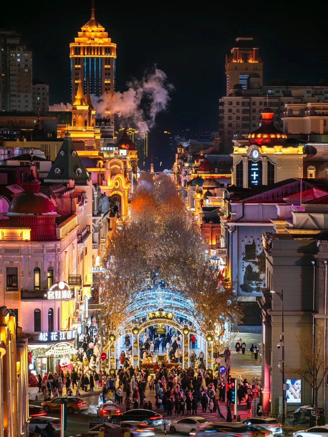 A nighttime view looking down a street lined with ornate, brightly lit buildings, a dense crowd of pedestrians walking under a large archway decorated with white lights, and cars driving on the street.