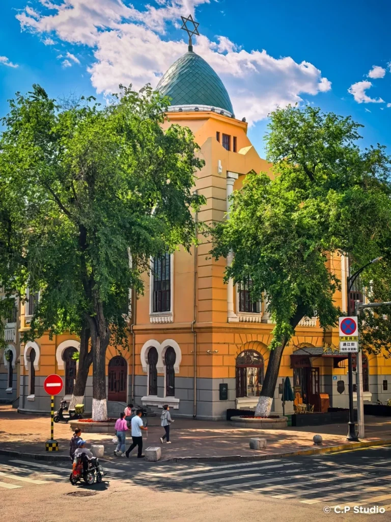 A yellow and grey building with arched windows and a green dome topped with a Star of David. Large trees with green leaves flank the building. Several people are walking on the sidewalk in front of the building. A crosswalk is visible in the foreground.