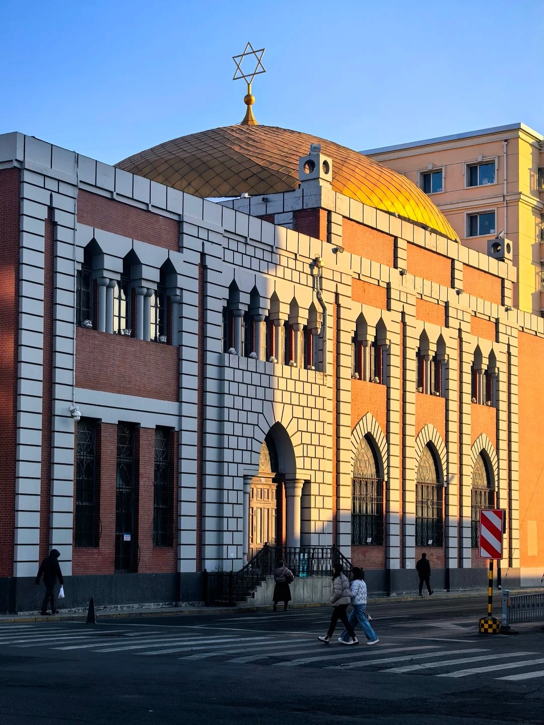The golden dome of the Harbin New Synagogue, topped with a Star of David, is visible above the brick and white-trimmed facade of the building. Several arched windows adorn the facade, and people are walking across a crosswalk in front of the building.