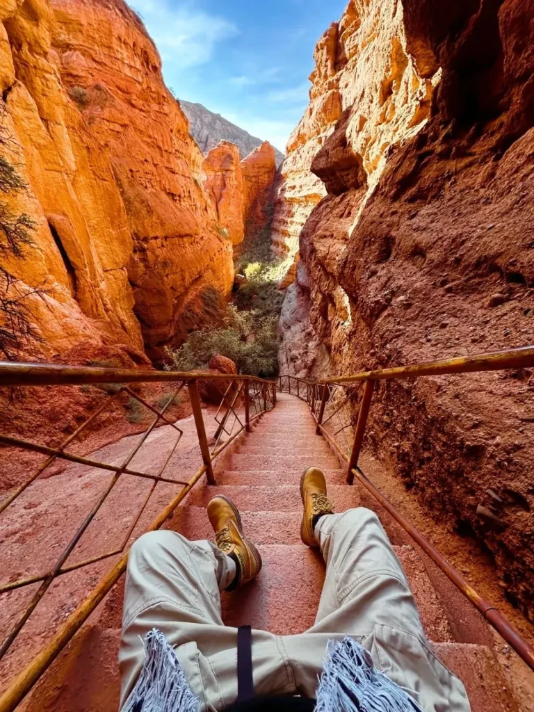 A person sitting on stone steps with tan boots and beige pants between tall red rock canyon walls with a metal handrail and blue sky above.