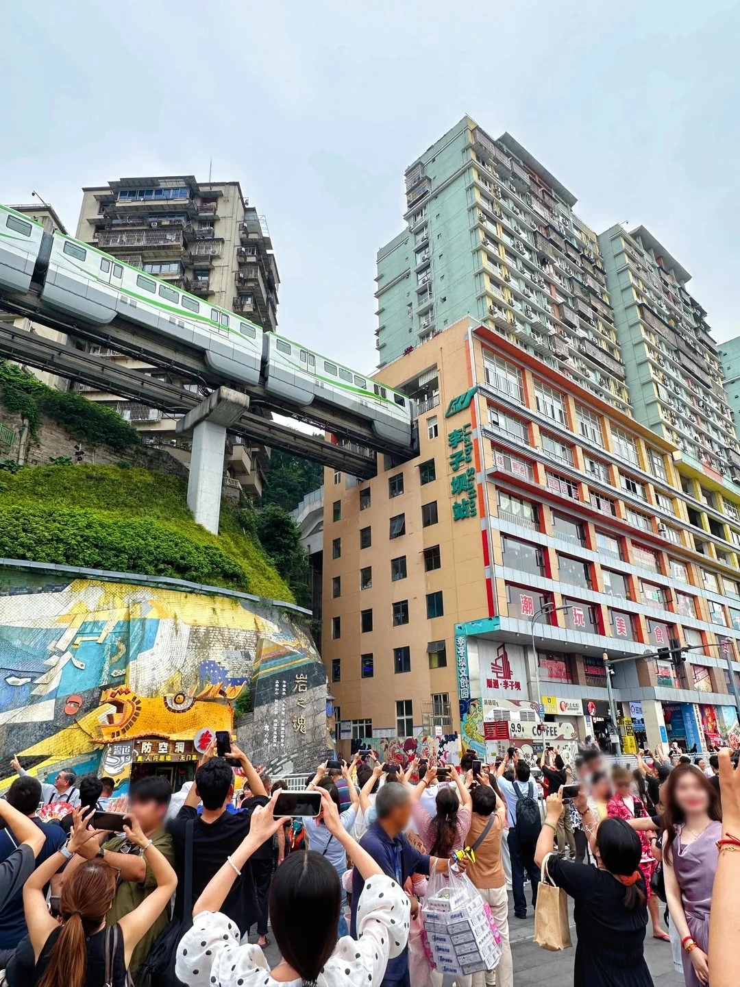 A monorail train passing between tall residential buildings above a crowded street with people taking photos in an urban city scene.