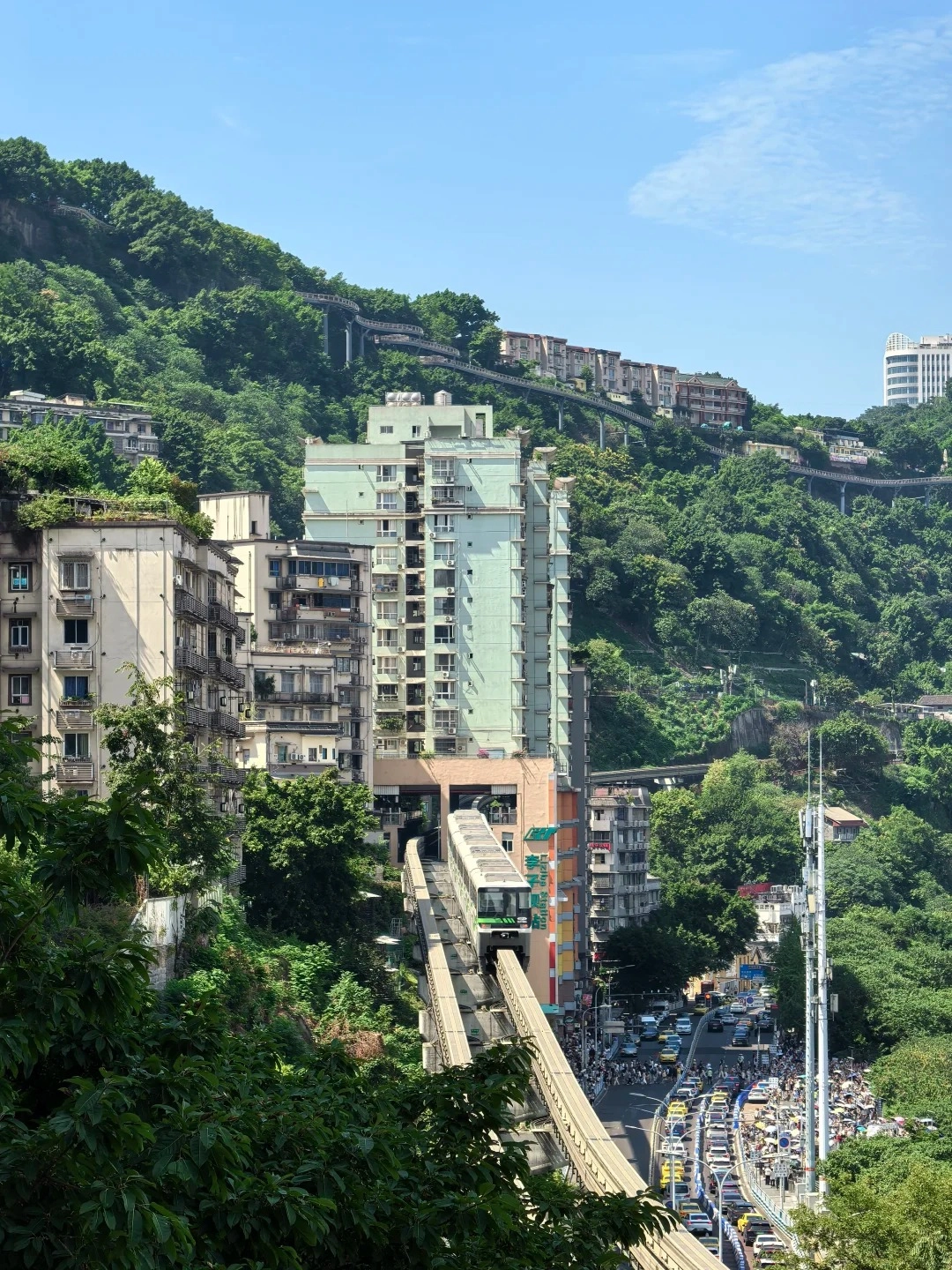 A funicular tram ascending a steep hillside beside tall residential buildings, dense green trees, and a busy city street below under a blue sky.