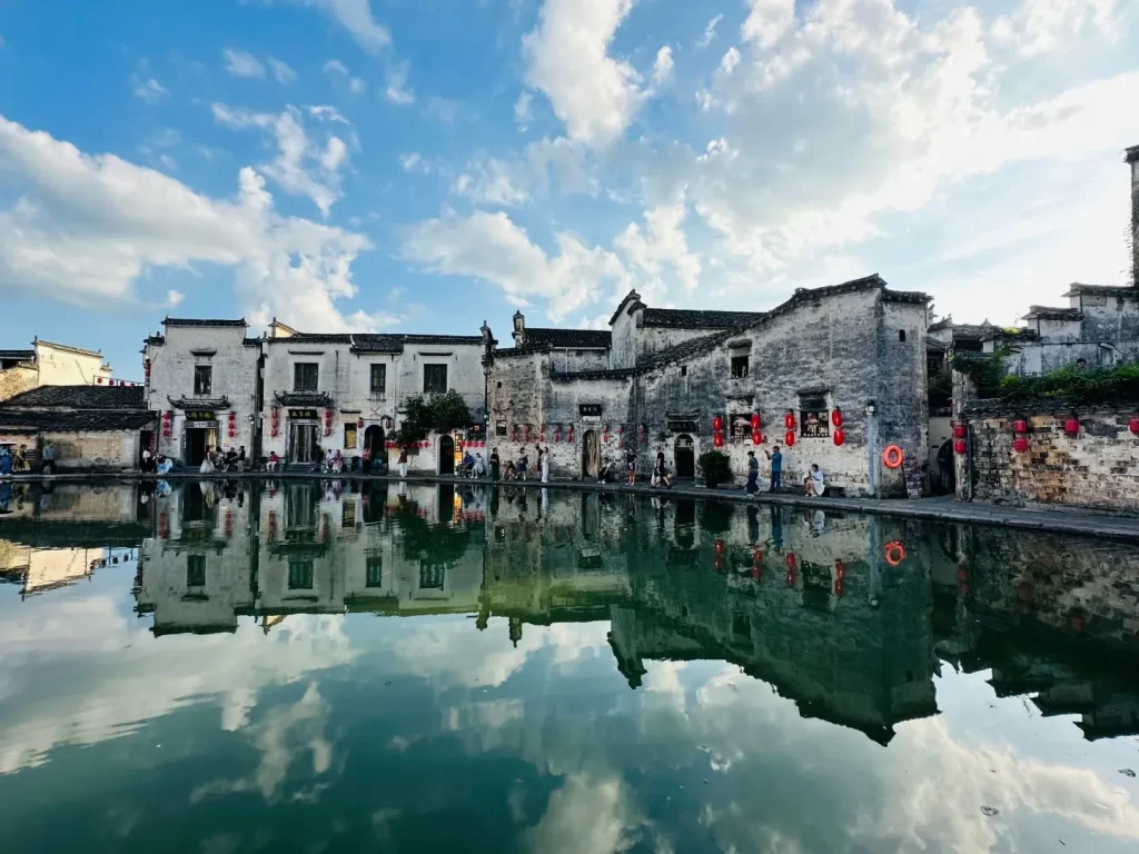 Whitewashed traditional Huizhou buildings with dark tiled roofs line the edge of a still pond, their reflections clearly visible in the water. Red lanterns hang from the buildings. The sky above is blue with scattered white clouds.