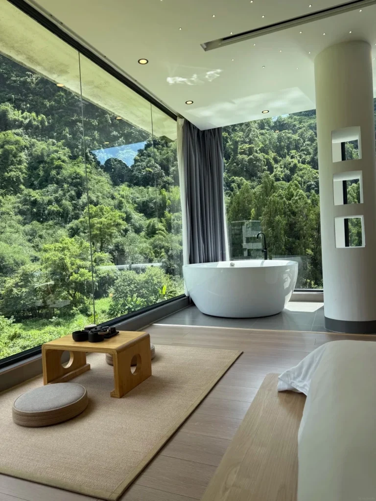 A modern hotel bathroom with a large freestanding white bathtub positioned in front of a floor-to-ceiling window offering a panoramic view of lush green mountains.