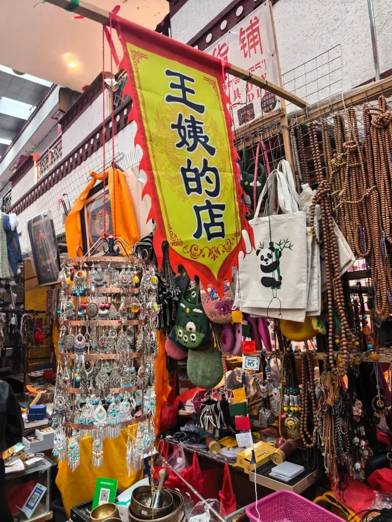 A vibrant market stall in Lhasa displays a large yellow banner with Chinese characters and a dragon illustration, hanging above racks of silver jewelry, felt bags, wooden beaded necklaces, and woven items. In the foreground, several brass singing bowls are visible.