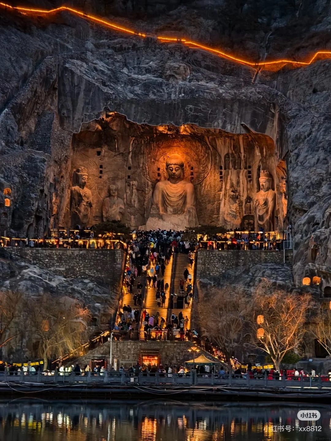 A wide-angle night view of the Longmen Grottoes, a UNESCO World Heritage site in Luoyang, China. Large, ancient Buddhist statues carved into a cliff face are illuminated by warm orange lights. A grand stone staircase filled with many tourists ascends towards the grottoes. The scene is set against a dark, rocky cliff and reflects on the calm water of the Yi River in the foreground. String lights with a vibrant orange glow line the top of the cliff.