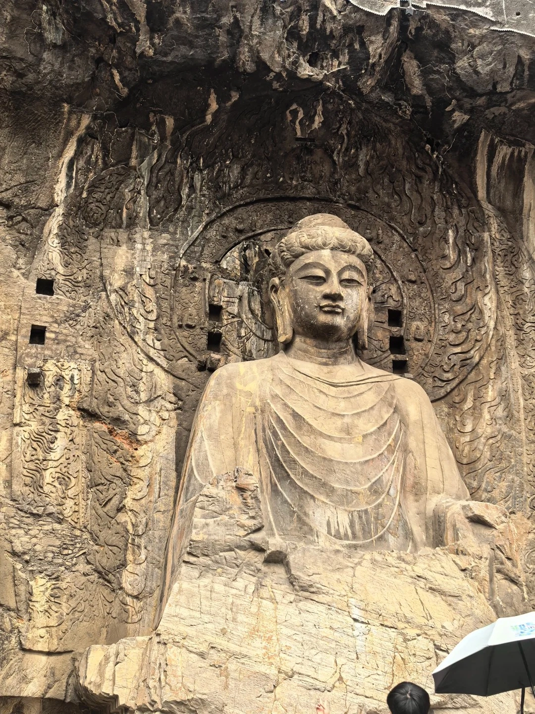 A large, stone-carved Buddha statue seated in a rocky alcove. The Buddha has a serene expression, with eyes downcast and lips slightly parted. It wears layered robes draped over its shoulders and chest. Behind the Buddha's head is a circular, carved halo with radiating patterns. The surrounding rock face is intricately carved with decorative motifs and has square indentations. The bottom of the image shows a hint of a dark umbrella and the head of a person looking up at the statue.