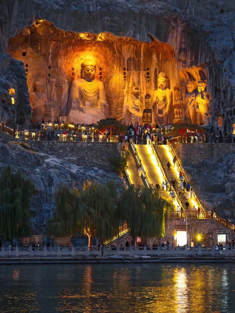 A colossal Buddha statue carved into a rocky cliff face, illuminated by warm lights at night. To the left of the main Buddha is a smaller Buddha statue. Below the statues, a wide stone staircase ascends the cliff, lined with lights and railings, with numerous people climbing. In front of the cliff base, there's a viewing area with railings and trees, and a body of water reflecting the lights.
