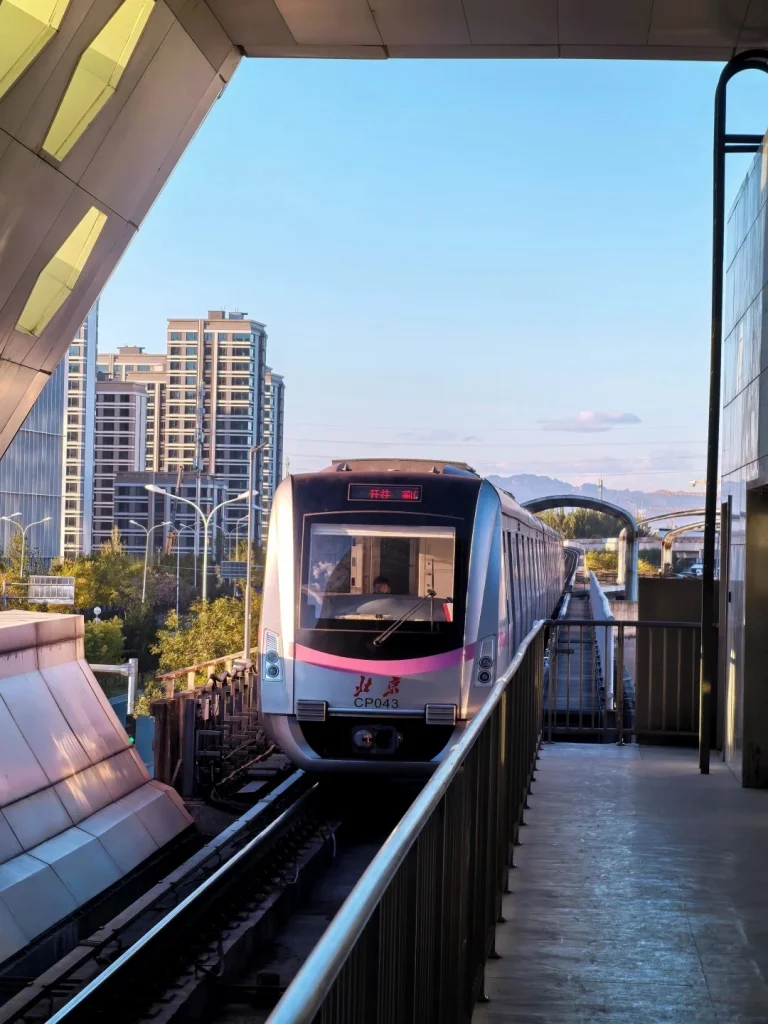 A monorail train approaching an elevated station track with city buildings, railings, and a clear blue sky in the background.