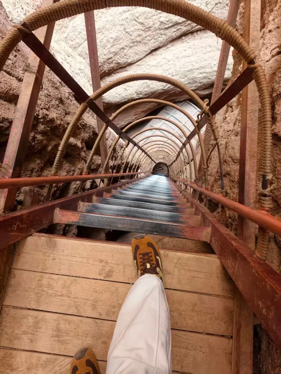 A steep narrow metal staircase with rope arches running through a rocky cliff tunnel, viewed from above with one person’s shoe visible on a wooden landing.