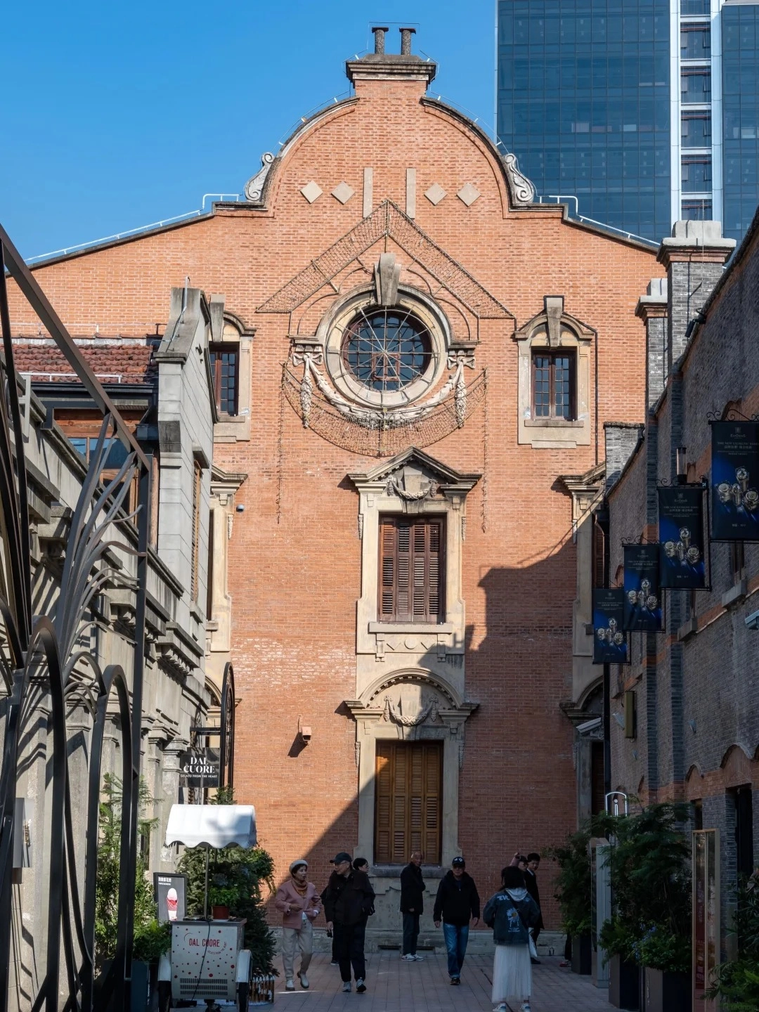A wide-angle shot of the ornate red brick facade of an old building in Shanghai. The building has a large, circular stained-glass window at the top, flanked by smaller rectangular windows. Below the circular window is a smaller rectangular window with wooden shutters, and beneath that is a large wooden door. The building is framed by narrower brick buildings on either side, with banners hanging from the right side. Several people are walking down the cobblestone street in front of the building, and a small ice cream cart is parked on the left.