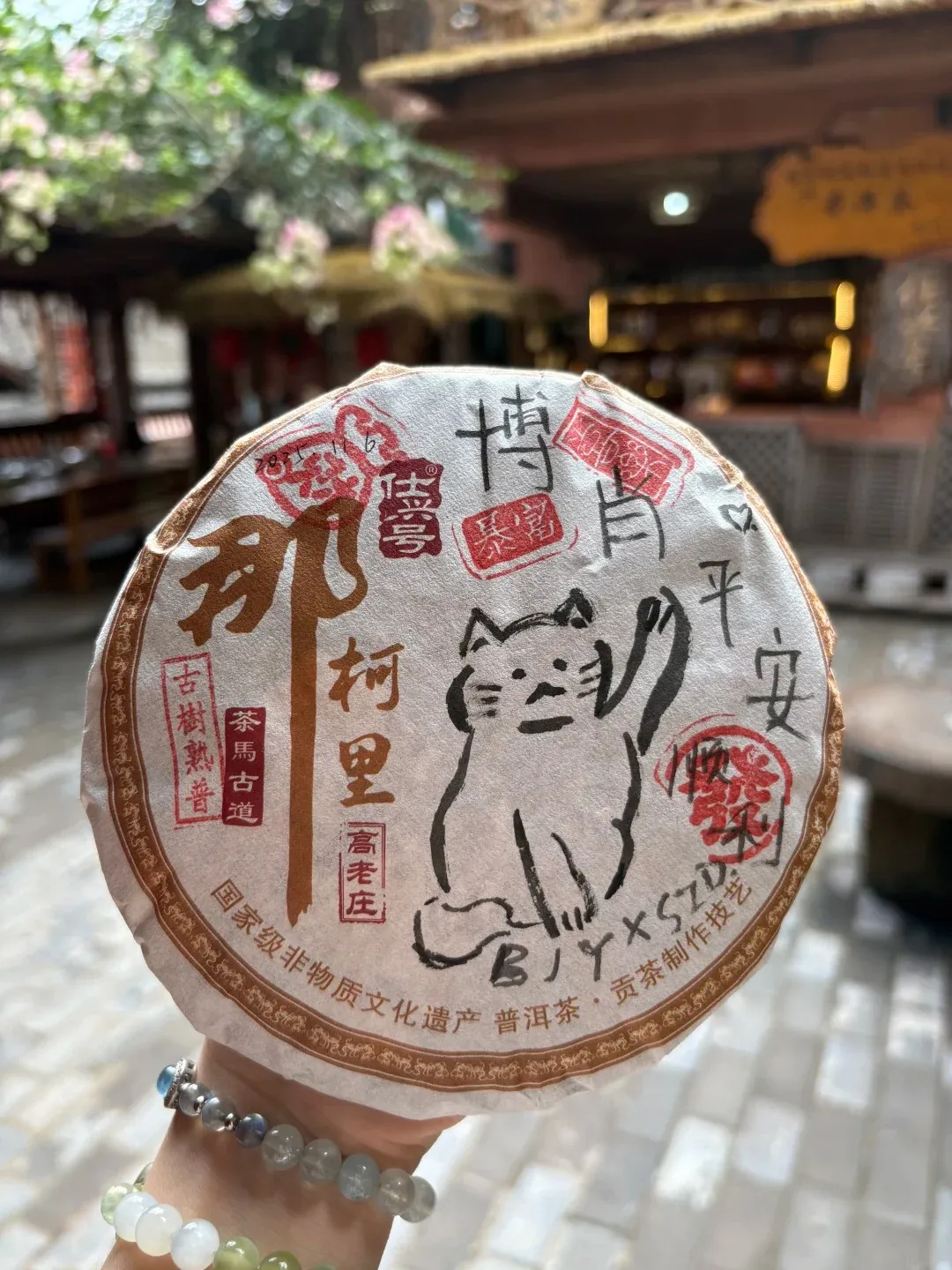 A round, compressed cake of Pu'er tea wrapped in paper, featuring a hand-drawn illustration of a cat with its paw raised, surrounded by Chinese calligraphy and red seals. The cake is held by a hand adorned with beaded bracelets.