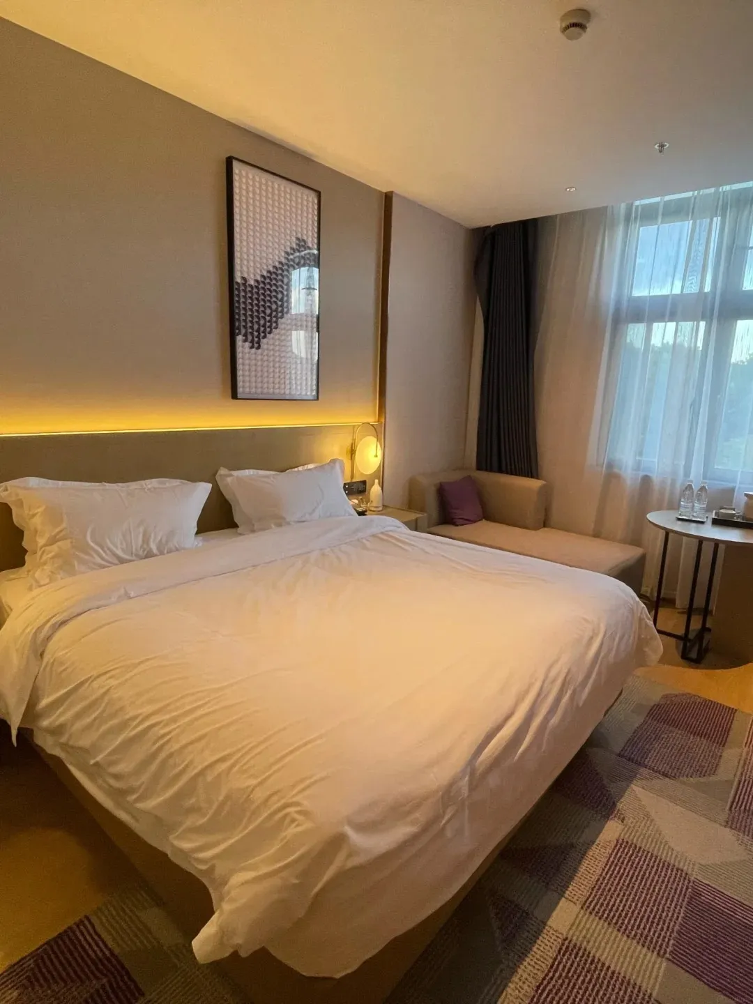 A hotel bedroom with a large, neatly made bed covered in white linens and pillows. A modern artwork hangs on the taupe wall above the headboard, which is illuminated by a strip of warm yellow light. To the right, a window with sheer white curtains reveals daylight, and a small seating area with a chaise lounge and a round table with chairs is visible.