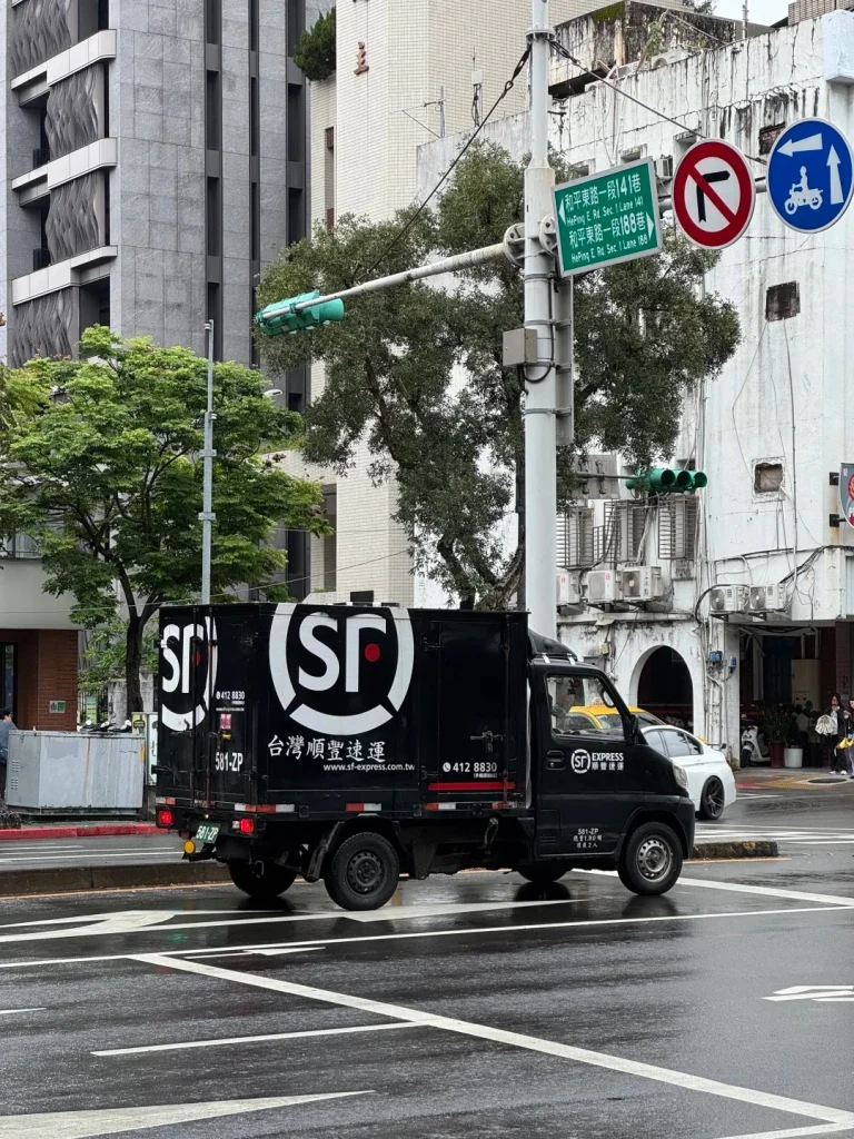 A black SF Express delivery truck is driving through a wet city intersection in Taiwan. The truck has large white SF Express logos and Chinese text reading '台灣順豐速運' (Taiwan SF Express). There are traffic lights, street signs with Chinese and English text, and buildings in the background. The road is wet and reflects the surrounding environment.