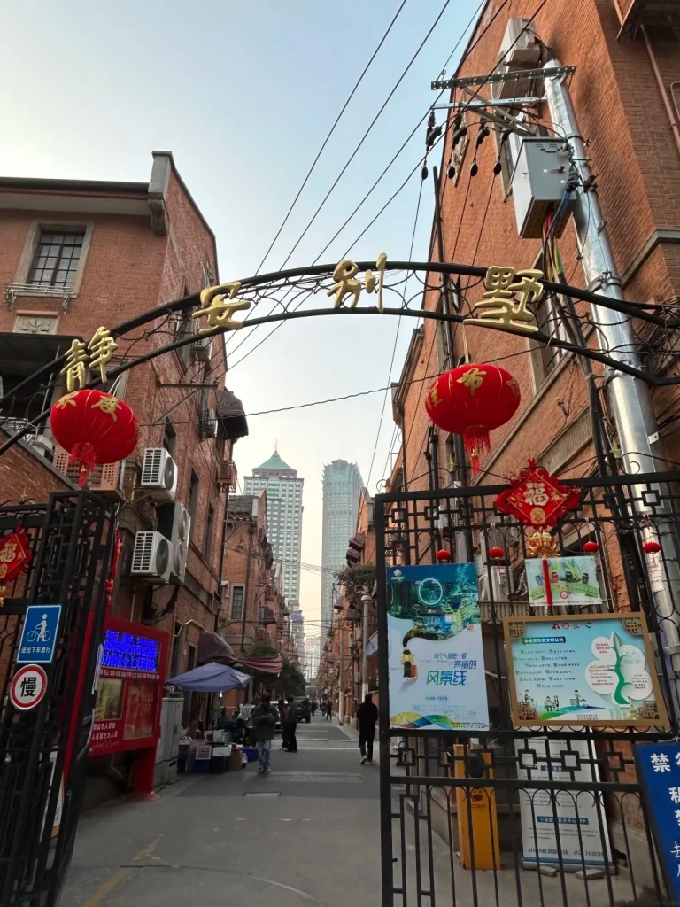 An ornate metal archway with gold Chinese characters spans the entrance to a narrow alleyway lined with red-brick buildings. Red lanterns hang from the archway and the buildings. A tall modern skyscraper is visible in the distance.
