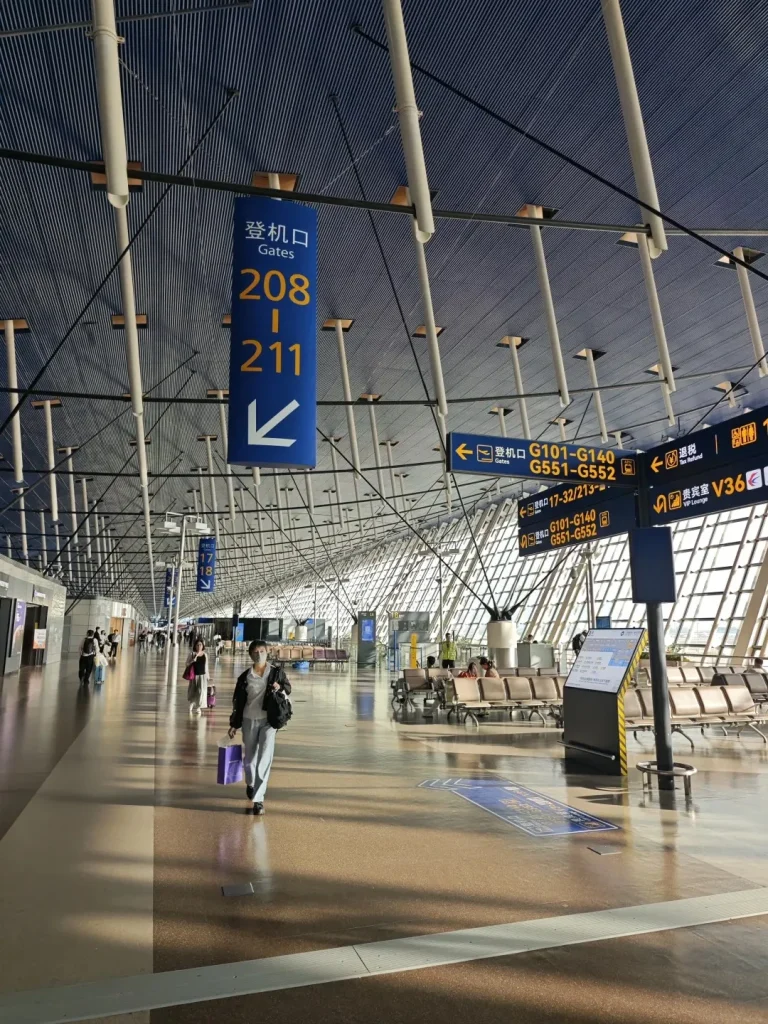 Interior view of a modern airport terminal with a high, arched ceiling constructed of metal beams and blue corrugated panels. Large white cylindrical columns support the ceiling structure. Overhead, blue directional signs are suspended, with one prominent sign indicating 'Gates 208-211' in Chinese and English, accompanied by a white arrow pointing left. Other yellow and blue directional signs for gates and amenities are visible further down the concourse. The floor is polished brown with light reflecting from the large, grid-patterned windows that form the terminal's facade. Several travelers are walking along the wide concourse, with a woman in the foreground wearing a white mask and carrying a purple shopping bag. Rows of waiting area seats are visible in the background near the windows.