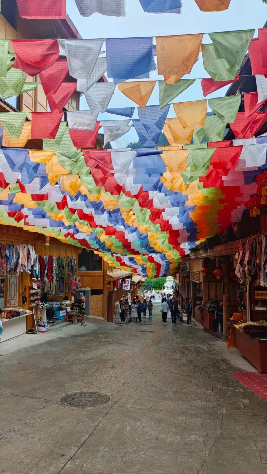 A narrow street lined with shops is covered overhead by numerous colorful Tibetan prayer flags strung across the sky.