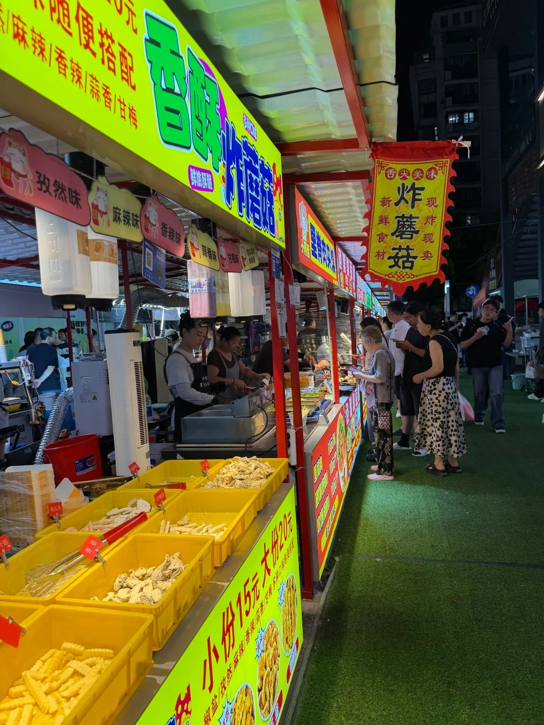 A brightly lit street food stall in China at night, with a selection of fried foods in yellow bins. Several people are standing at the counter, and a banner with Chinese characters hangs above. The stall has a green turf-like ground covering in front.