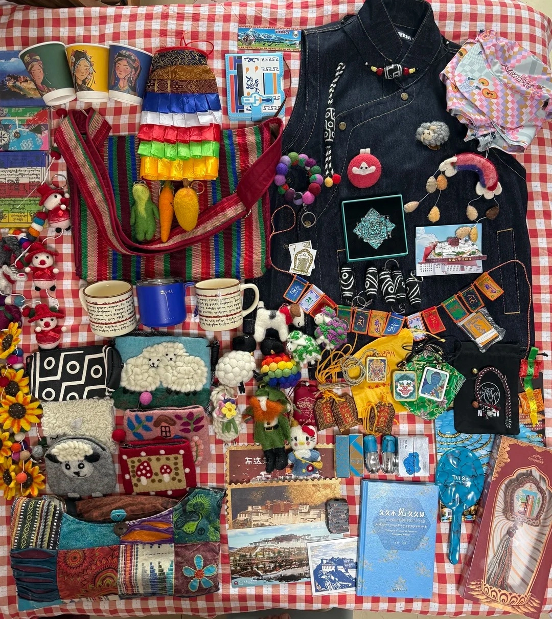 A flat lay of various Tibetan souvenirs arranged on a red and white checkered tablecloth. Items include felt ornaments shaped like sheep and clouds, small embroidered pouches, a denim vest with decorative stitching, colorful beaded bracelets, small flags with Tibetan script, cups, and books.