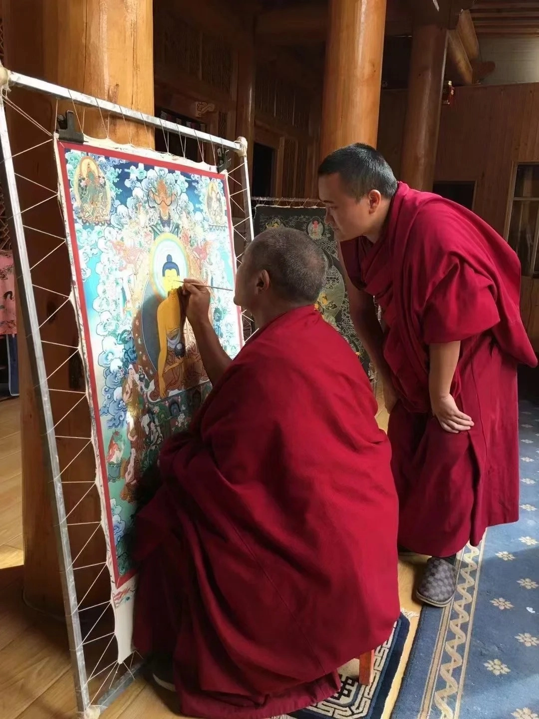 Two Buddhist monks in red robes are painting a colorful thangka on a frame. The older monk in the foreground is kneeling and meticulously painting the face of a central Buddha figure with a fine brush. The younger monk stands behind him, looking closely at the artwork. The thangka depicts various deities and celestial scenes against a vibrant blue background with clouds.