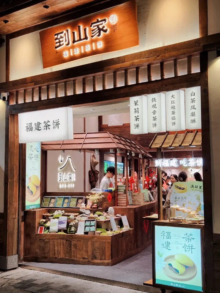 A storefront of a traditional Chinese tea shop with wooden architecture. A prominent wooden sign with Chinese characters is illuminated above the entrance. Below the sign are hanging lanterns with Chinese text advertising different teas. Inside, a display counter showcases various tea cakes and pastries, with a person serving customers in the background. A lit sign with an image of green pastries and Chinese text is visible in the foreground.