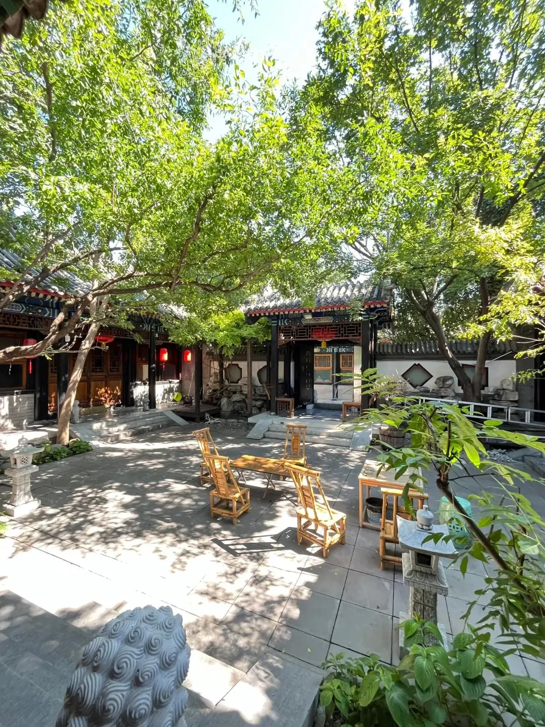 An open-air courtyard in a traditional Chinese siheyuan, shaded by lush green trees with dappled sunlight on the grey paved ground. Traditional Chinese architecture with dark wood beams and red accents surrounds the courtyard. In the center, there is a wooden dining table with several bamboo chairs. To the right, a stone lantern stands near potted plants and another small wooden table.