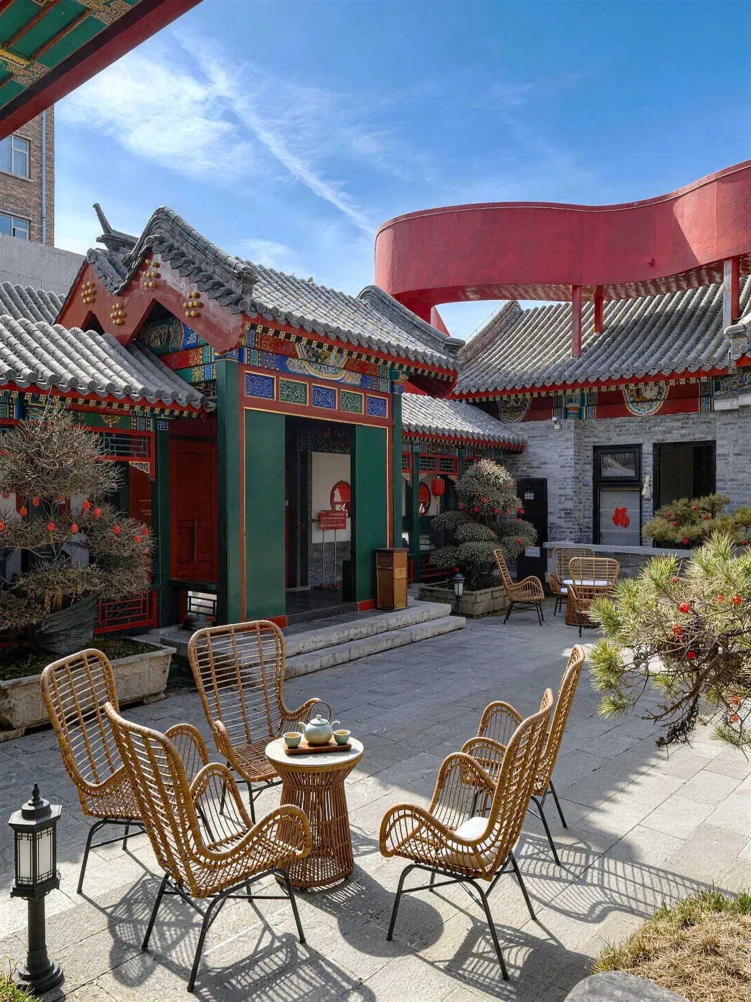 An outdoor courtyard with traditional Chinese architecture, featuring grey tiled roofs with ornate eaves, painted beams and columns in red and green. In the foreground, there are several rattan chairs surrounding a small, round table with a tea set. In the background, a modern red curved structure is visible above the traditional buildings.