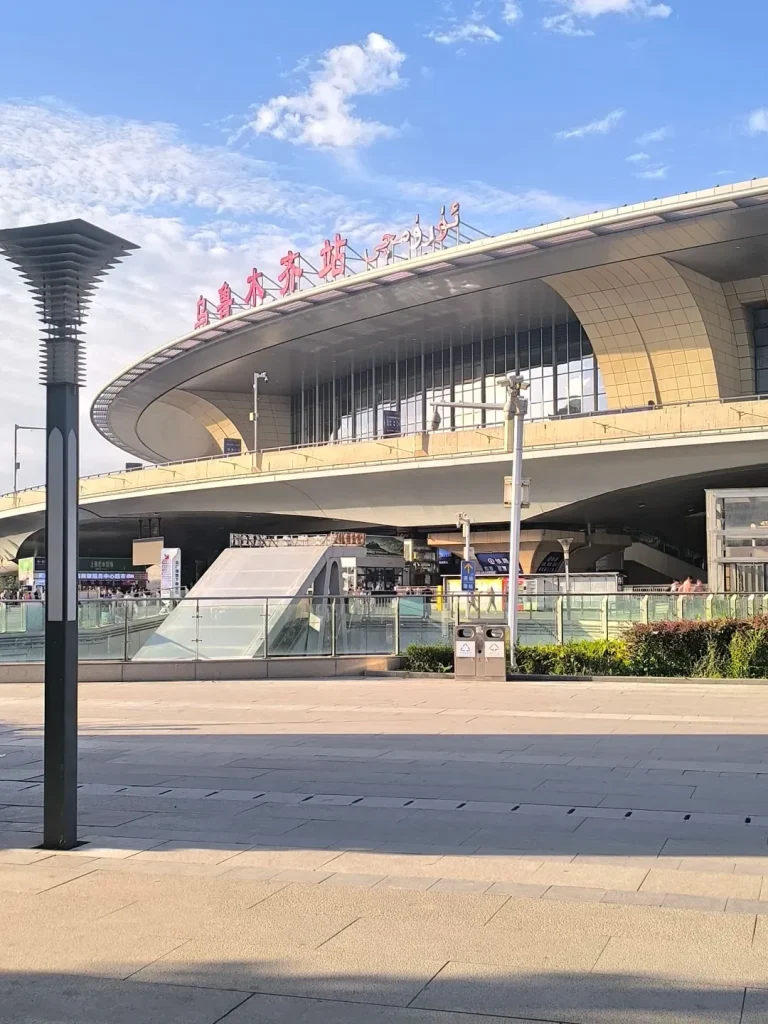 A modern train station building with a curved, beige, tiled roof and large glass windows. Red Chinese characters are visible on the roof's overhang. In the foreground, a tall, dark gray lamppost stands on a paved plaza. The sky above is blue with scattered white clouds.