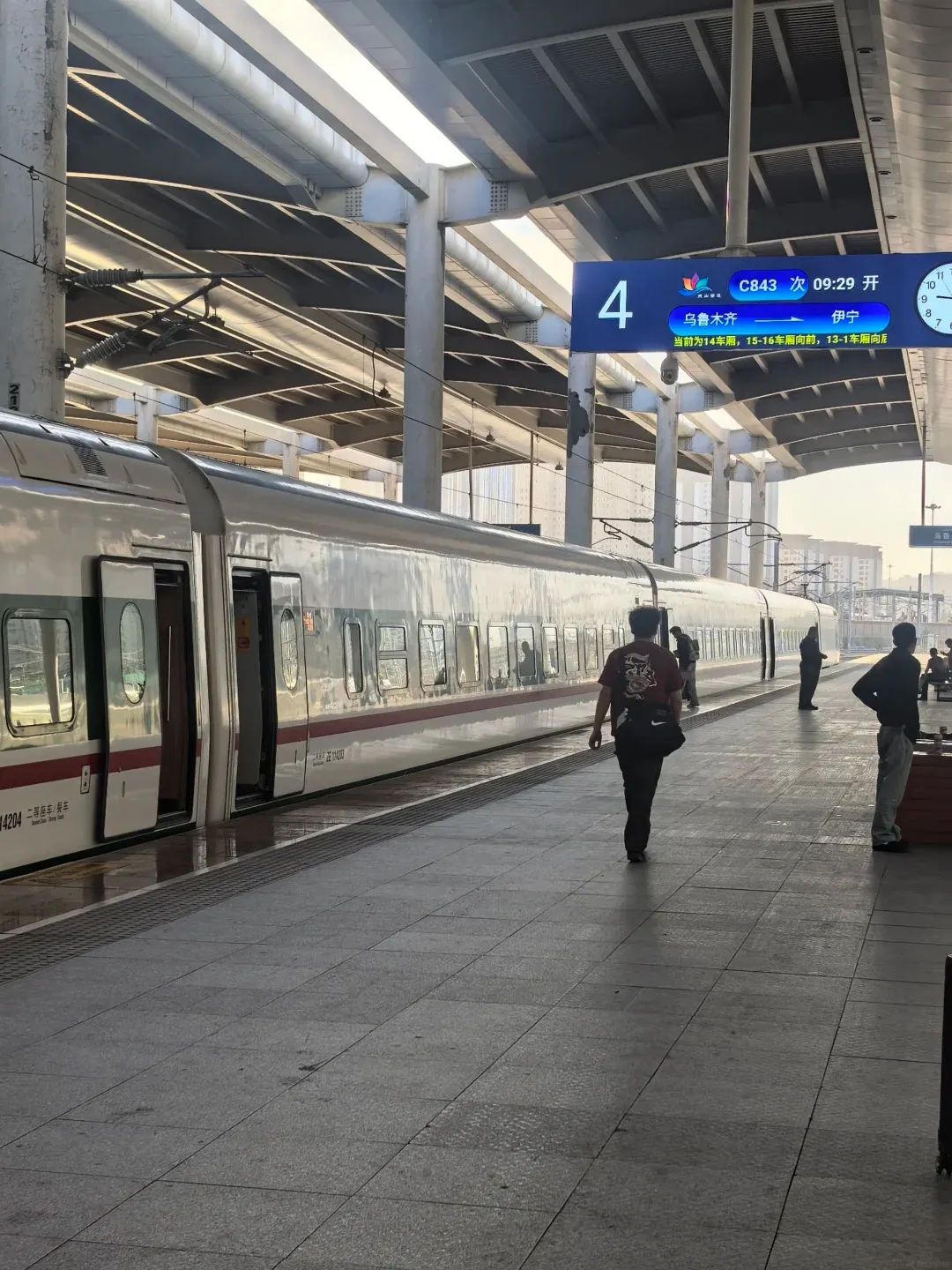A view from a train platform at Urumqi Railway Station in Xinjiang, China. A modern, white high-speed train with red and grey accents is stopped at the platform. Several people are standing on the platform, some walking. An electronic display board above shows train information including 'C843 09:29 开' and '4' for platform number. The platform is paved with large grey tiles, and a covered walkway with supporting pillars is overhead. Sunlight streams in from the side, casting shadows.