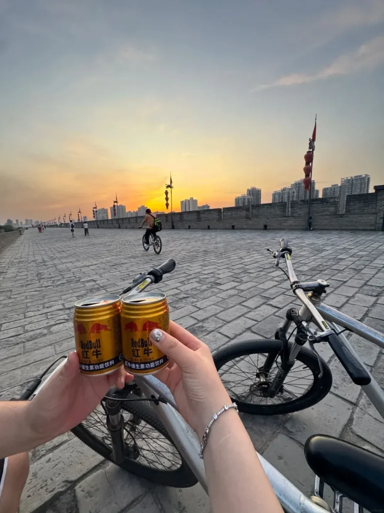 Two hands hold up two cans of Red Bull in front of two bicycles on the Xi'an City Wall during sunset, with people cycling and walking in the background and city buildings visible beyond the wall.