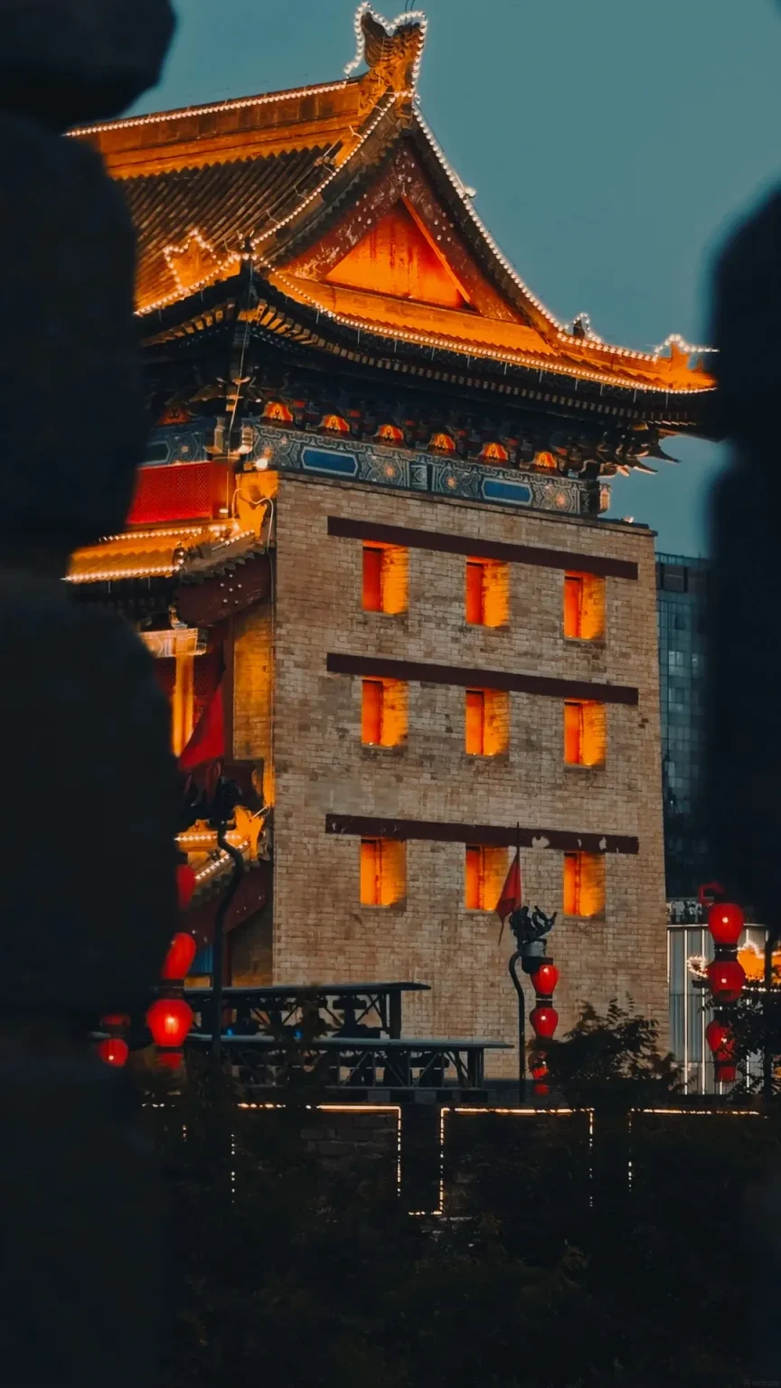 A traditional Chinese gate tower on the Xi'an City Wall is illuminated at dusk with orange lights highlighting its architecture and numerous red lanterns hanging around it.