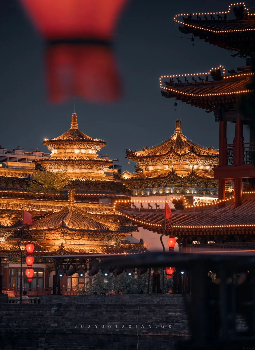 A night view of the illuminated Xi'an City Wall South Gate, with traditional Chinese architecture featuring tiered roofs adorned with lights, red lanterns hanging, and a dark sky.