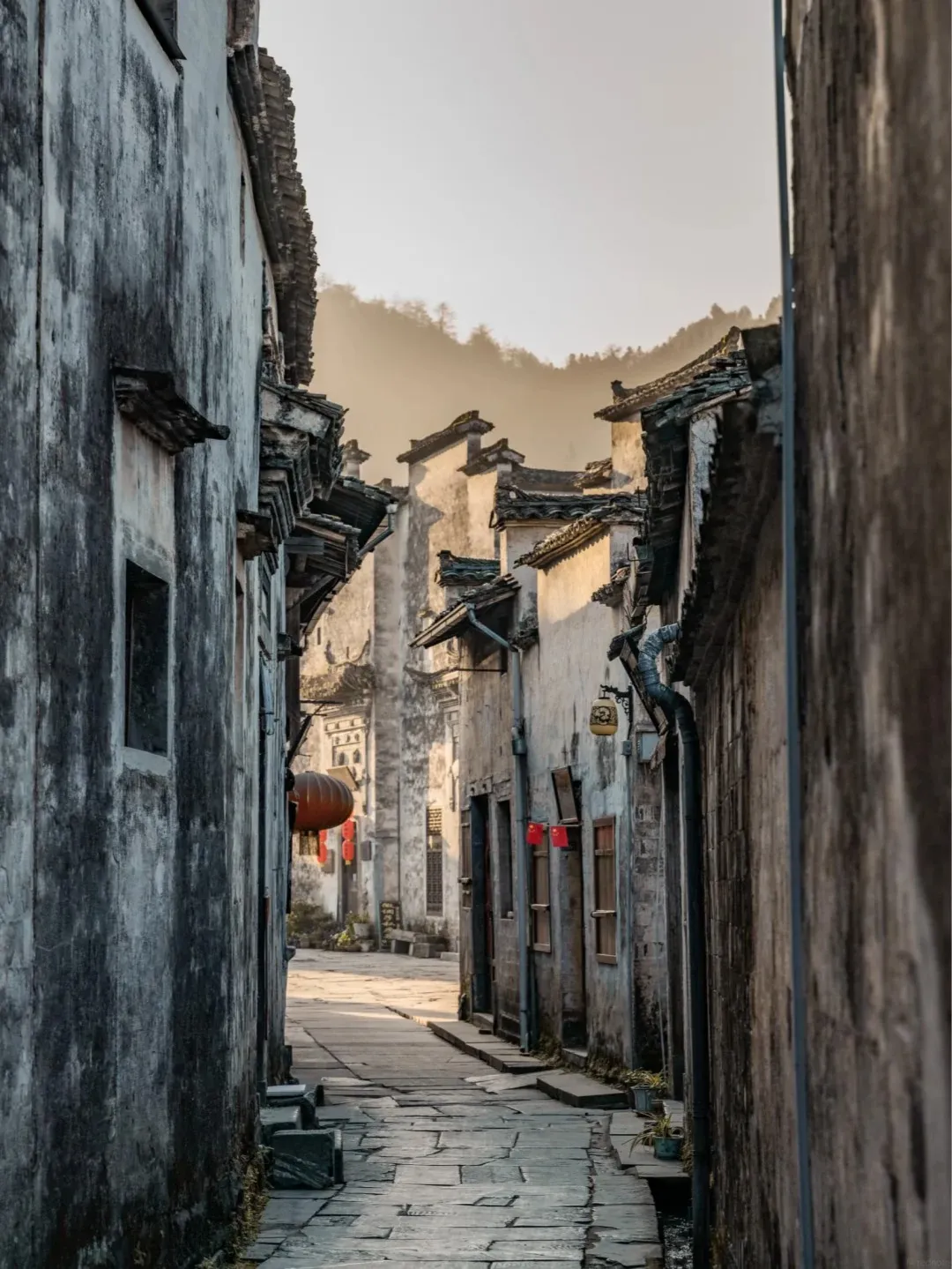 A narrow stone-paved alleyway in an ancient village, flanked by traditional white-walled buildings with dark tiled roofs. Red lanterns hang from the buildings, and a hazy, tree-covered hillside is visible in the background.
