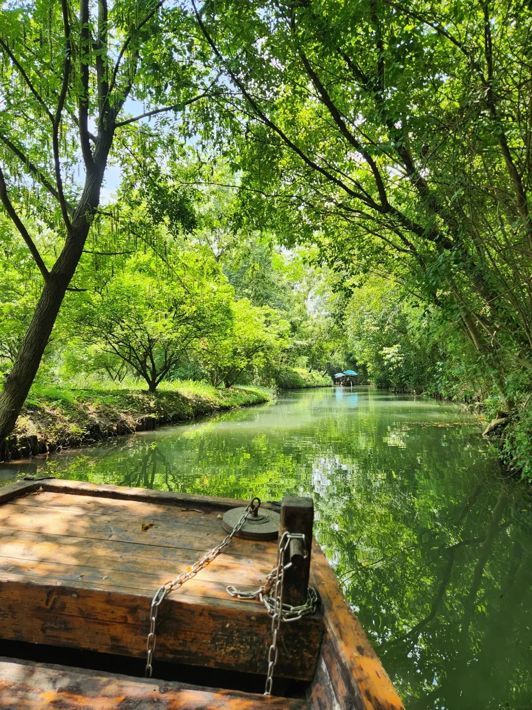 View from the front of a wooden boat with metal chains on its deck, looking down a narrow green canal lined with lush trees. The water reflects the green foliage and sky. In the distance, two blue umbrellas are visible on the water.