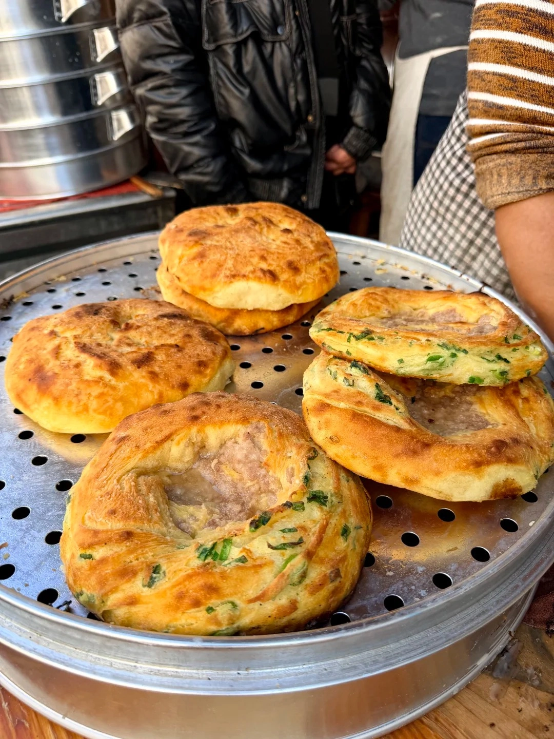 A close-up shot of several golden-brown, round flatbreads with a hole in the center, some filled with green onions and meat, arranged on a perforated metal tray.