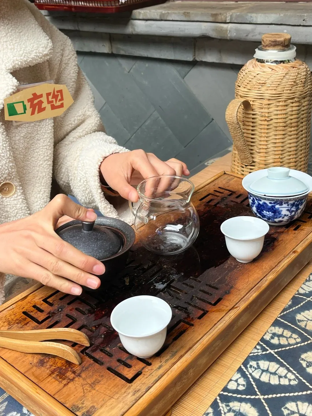 A person's hands are shown performing a tea ceremony on a wooden tea tray. There are several white teacups, a glass teapot, and a dark lidded teacup. A woven thermos is in the background.