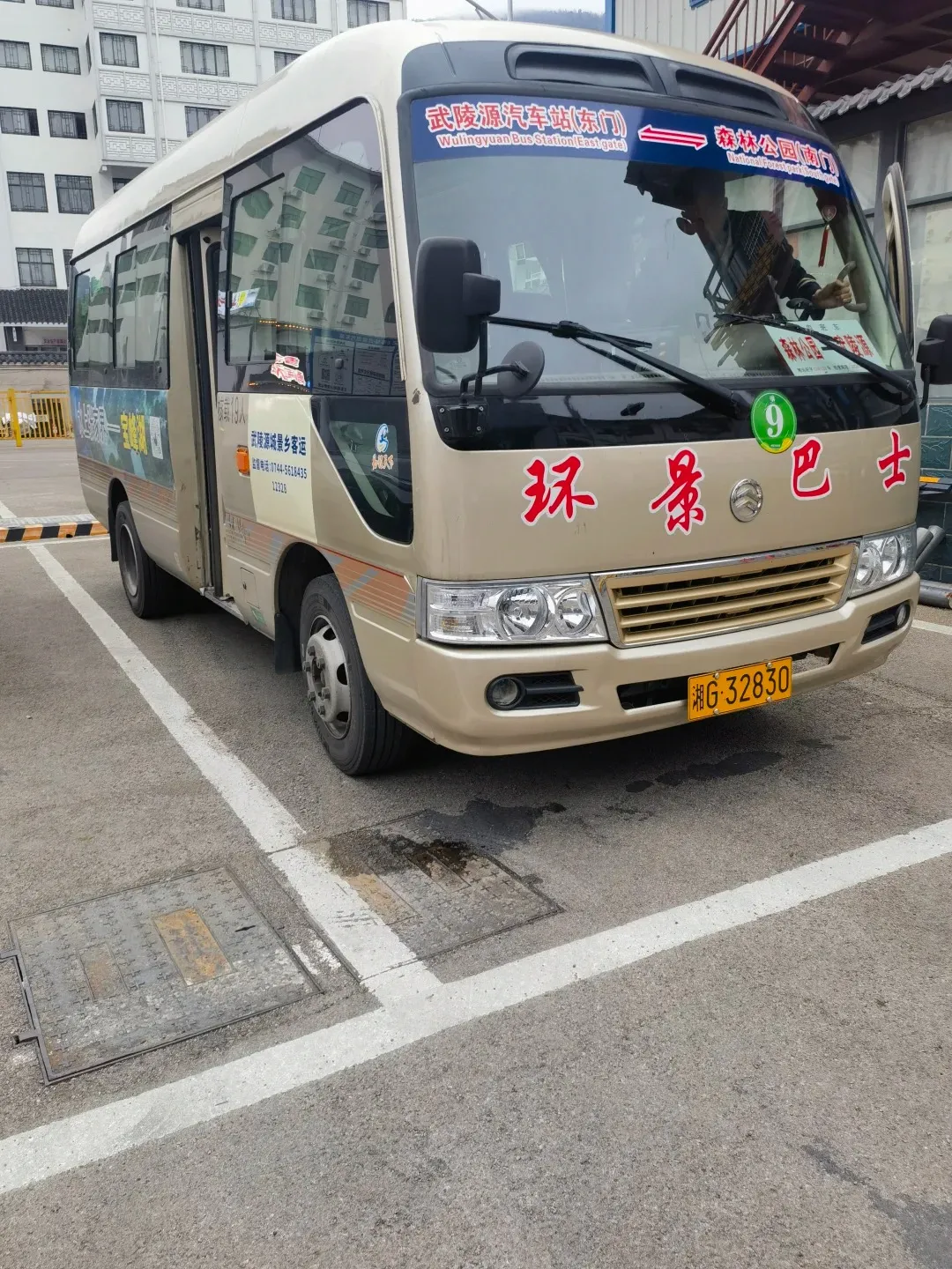 A gold and white shuttle bus parked in a lot, with Chinese characters and English text on its windshield. The text indicates it is a bus to Wulingyuan Bus Station (East Gate). The bus has large side windows and a side door open. A yellow license plate with Chinese characters and the number 'G-32830' is visible on the front.