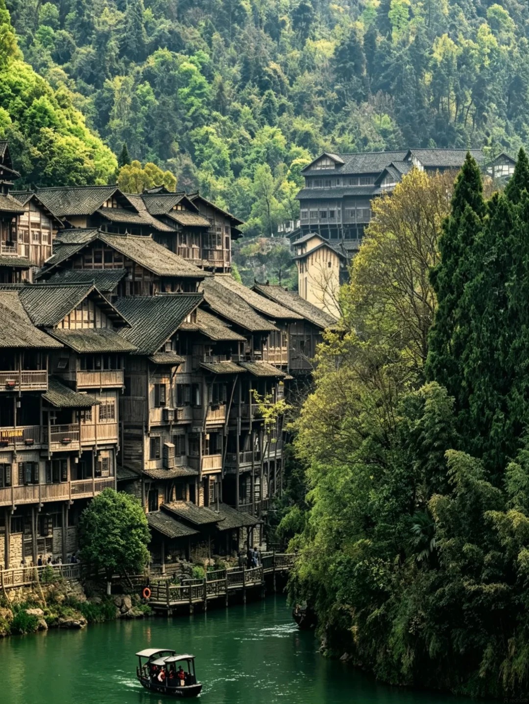 A scenic view of traditional wooden stilt houses with dark tiled roofs, built along a green river. A small boat with people on board navigates the river in the foreground. The village is nestled at the base of lush, tree-covered mountains.