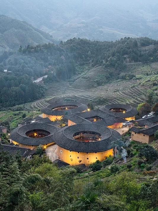 An aerial view of multiple large, circular, earthen Fujian Tulou buildings with dark tiled roofs, illuminated by warm lights, nestled in a verdant, mountainous landscape with terraced fields and scattered trees.