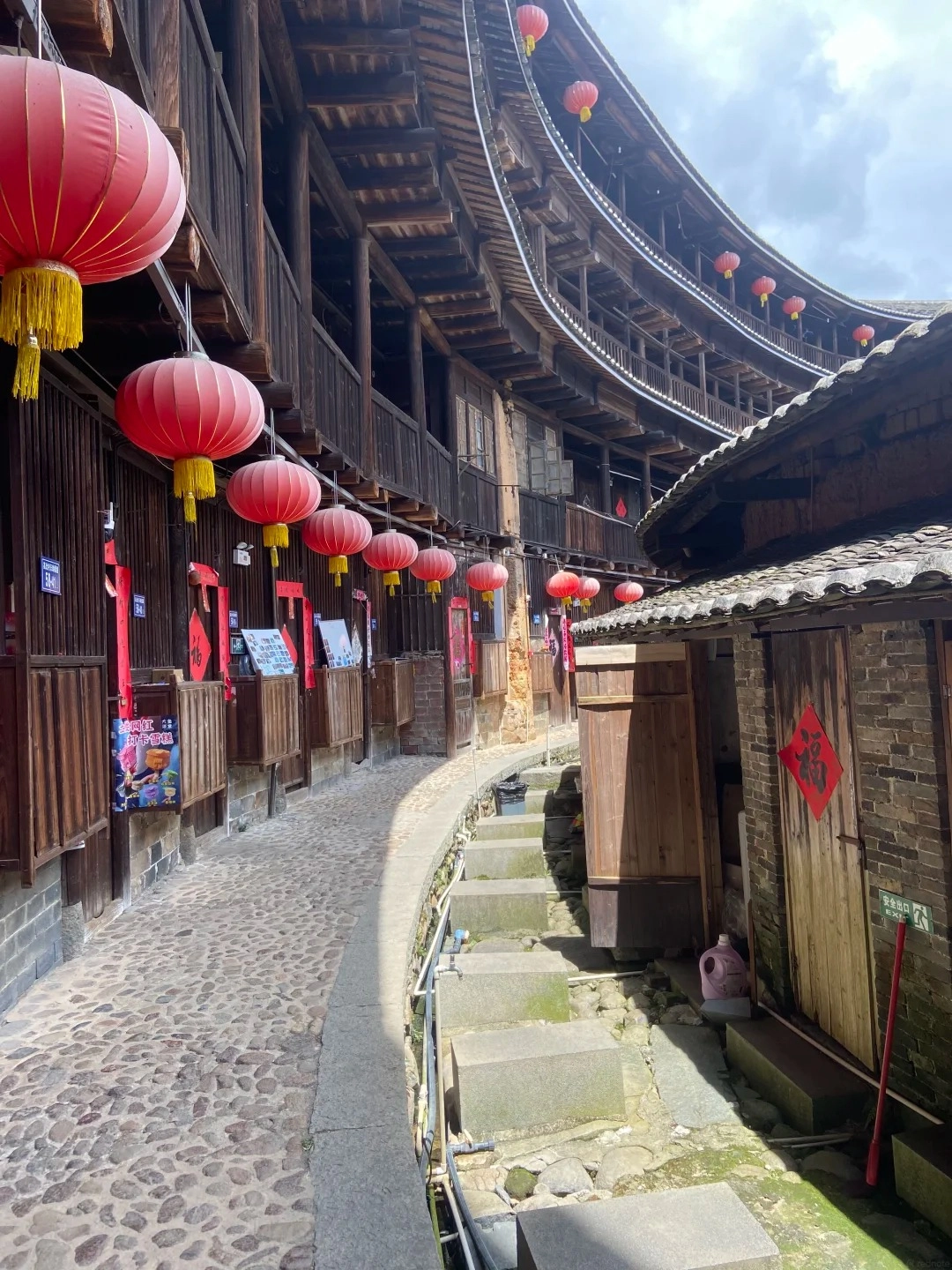 A curved, multi-story wooden building with open balconies and red lanterns hanging from the eaves, lining a cobblestone pathway. A narrow stone staircase descends to a lower level with wooden doors and brick walls.