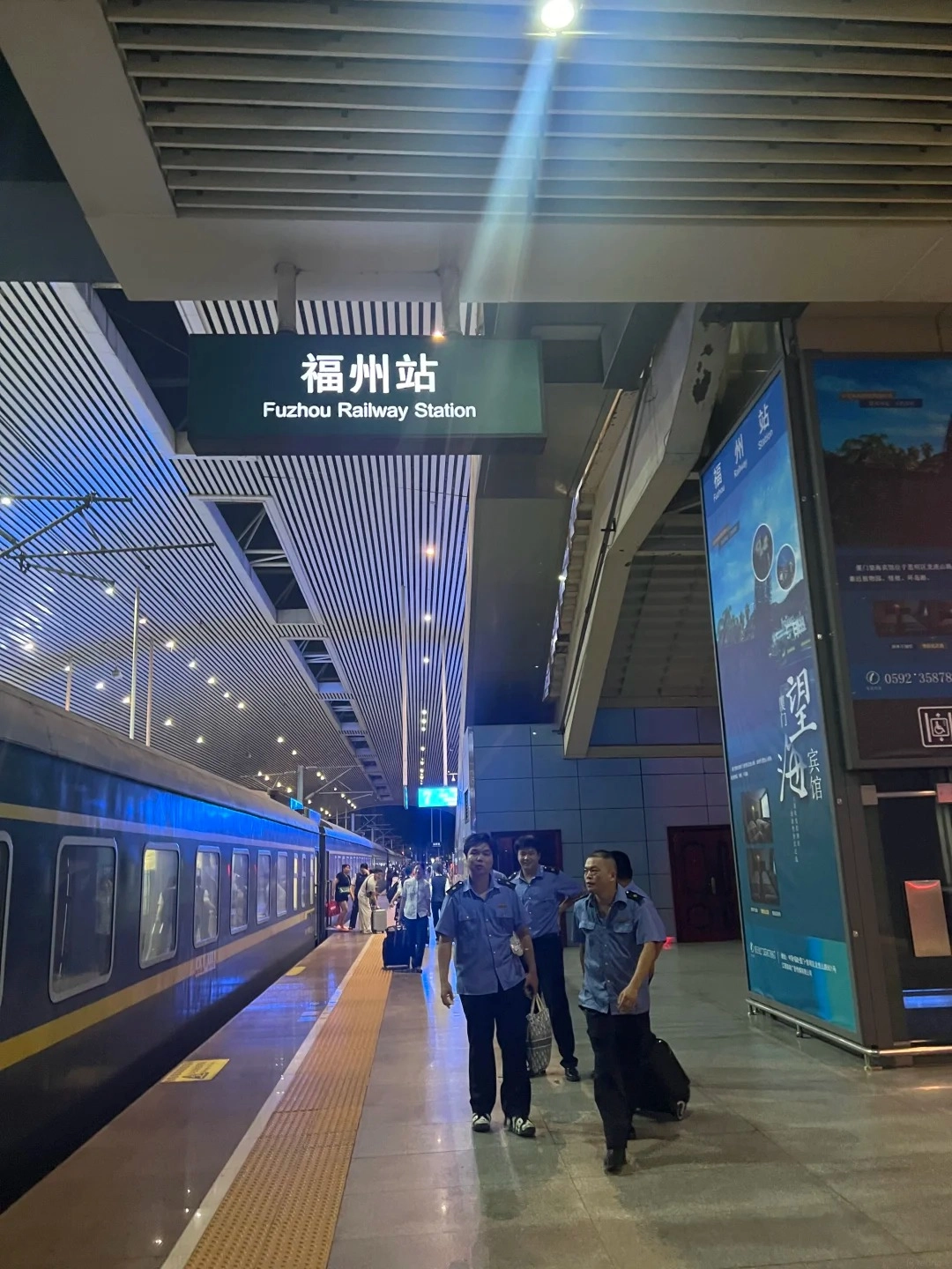 A train station platform at night with a train parked on the tracks. Several people, including uniformed staff, are walking on the platform. A sign overhead reads '福州站 Fuzhou Railway Station'. The train has blue and yellow accents and lit windows.