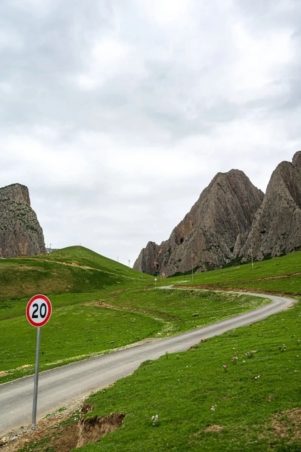 A winding paved road curves through lush green grassy hills under a cloudy sky. In the foreground, a round red and white speed limit sign shows '20'. In the background, imposing, rugged rock formations rise dramatically on either side of the landscape. There are power poles visible on the left and right sides of the road.