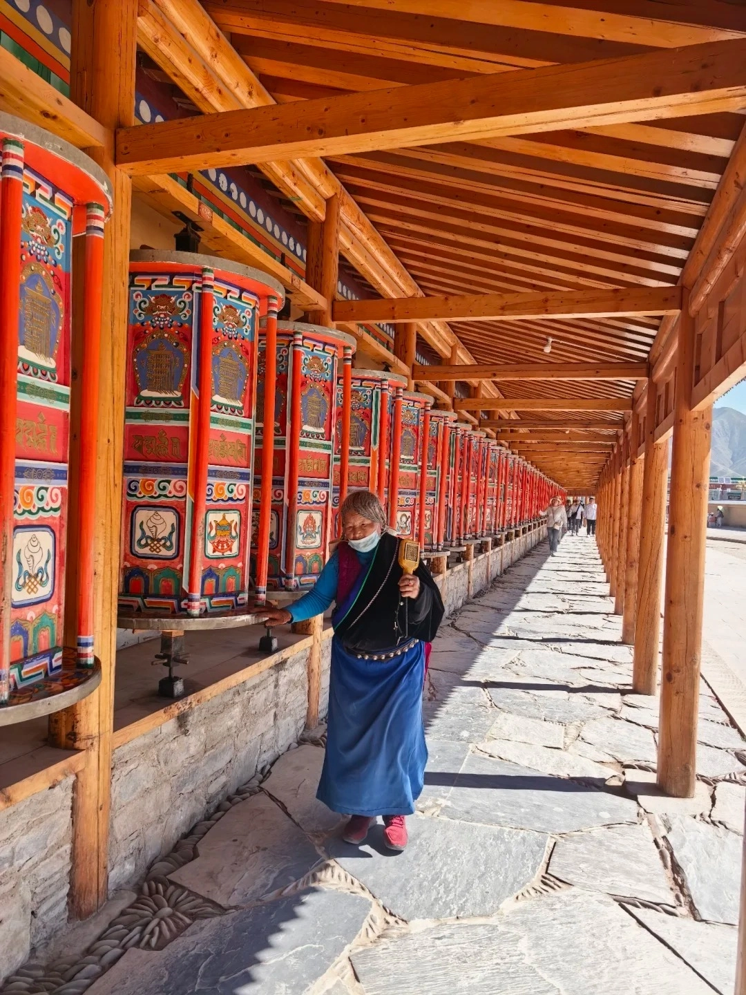 An elderly Tibetan woman wearing a face mask, blue skirt, and black jacket, is spinning a large, ornate, red and blue prayer wheel along a covered walkway lined with many other prayer wheels.