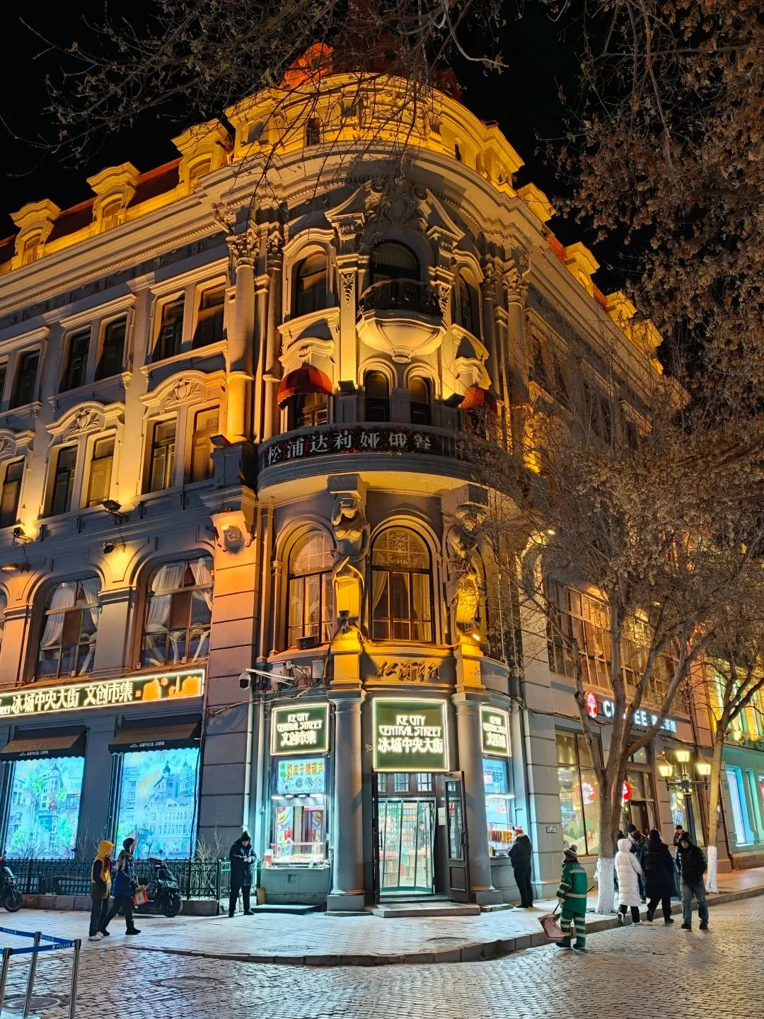 A corner view of a multi-story European-style building illuminated at night, with a domed roof section and ornate facade. The ground floor has shops with glowing signs, including one that says 'KC CITY CENTRAL STREET'. People are walking on the cobblestone street in front of the shops, and bare tree branches are visible against the dark sky.